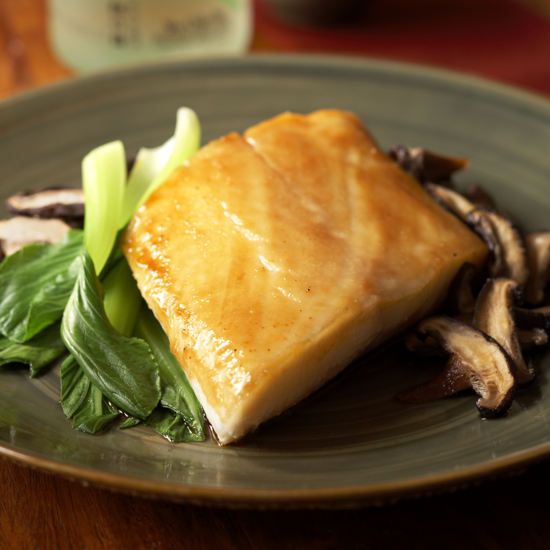 Plated black cod with greens and mushrooms on a wooden table.