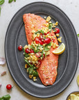 Cooked aalmon fillet with corn and tomato salad on a gray plate, surrounded by basil leaves and lemon wedges.s