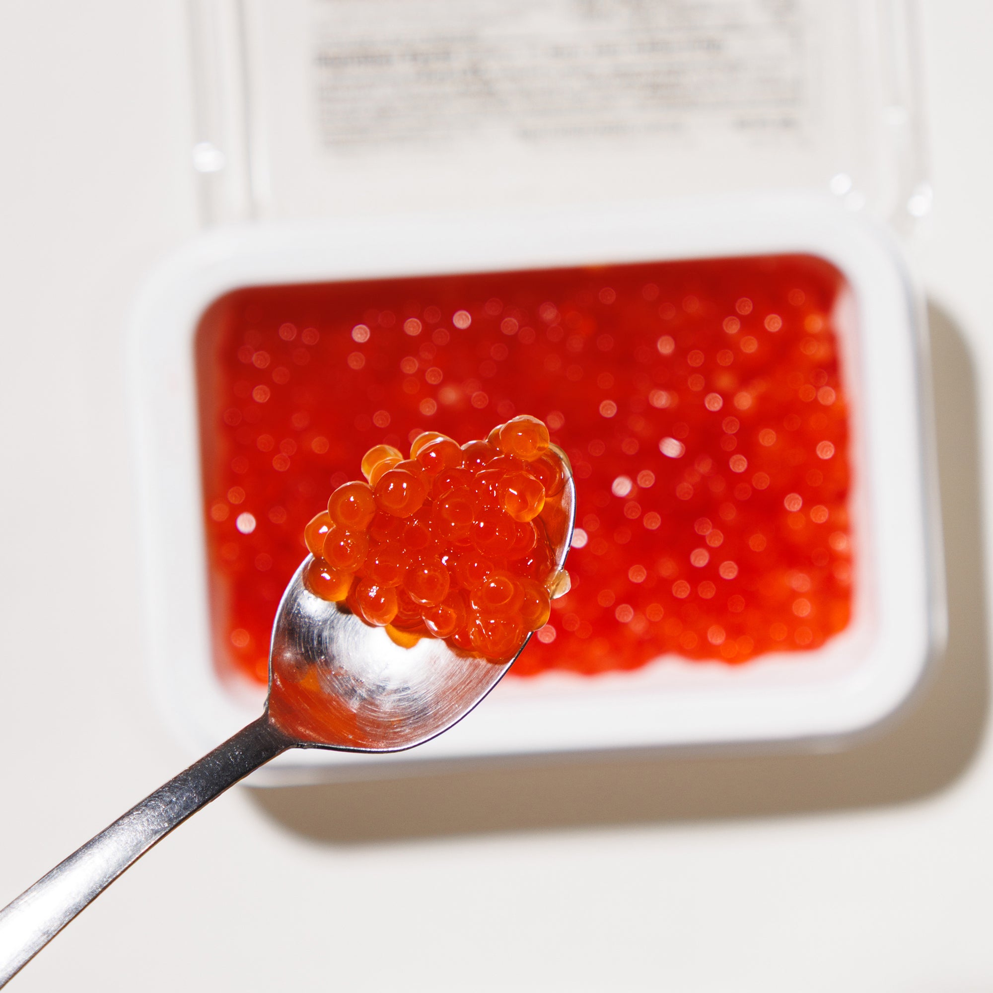 Spoonful of red caviar over a tray of caviar on a white background