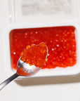 Spoonful of red caviar over a tray of caviar on a white background