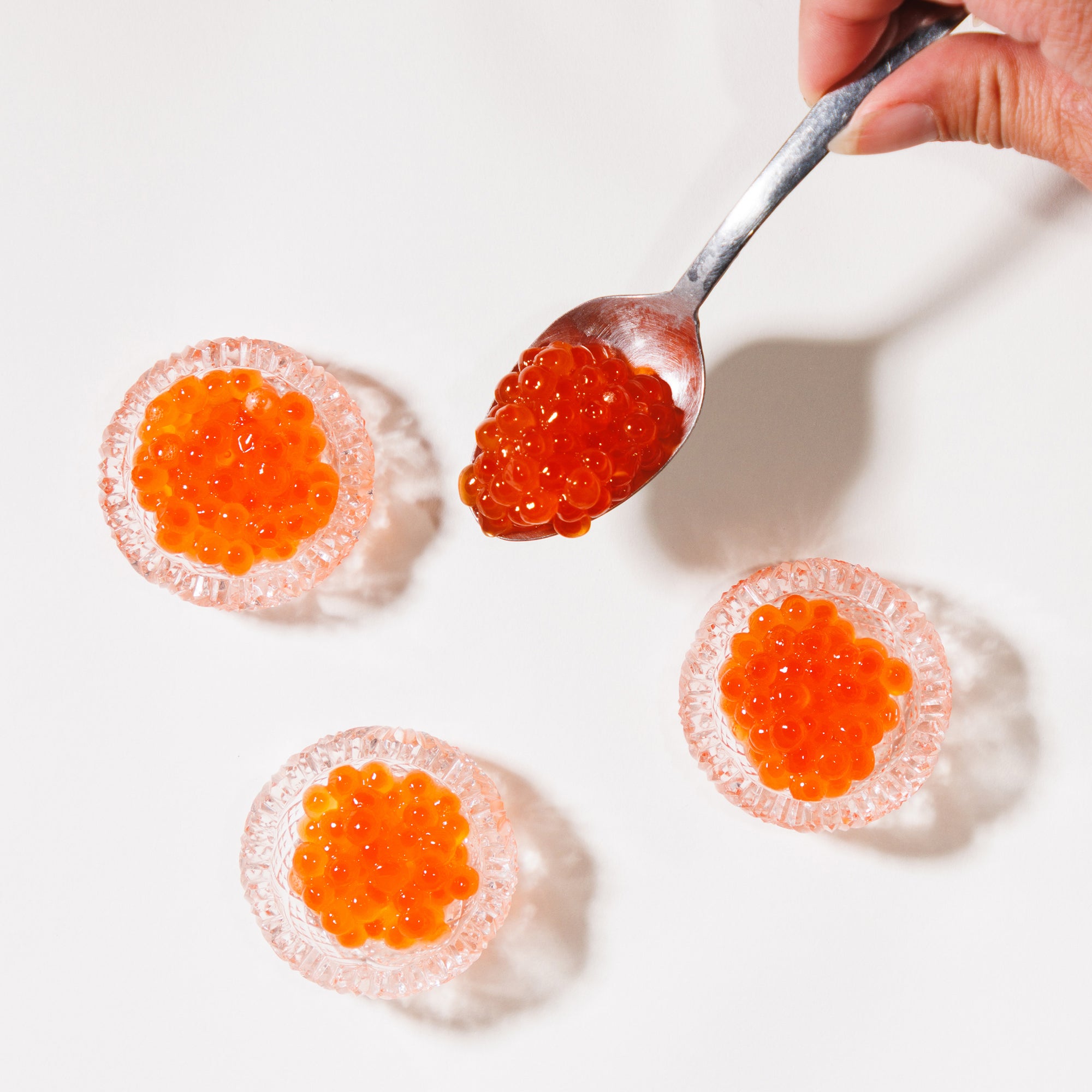 Spoonful of red caviar being added to three round clear mini glasses on a white background.