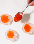 Spoonful of red caviar being added to three round clear mini glasses on a white background.