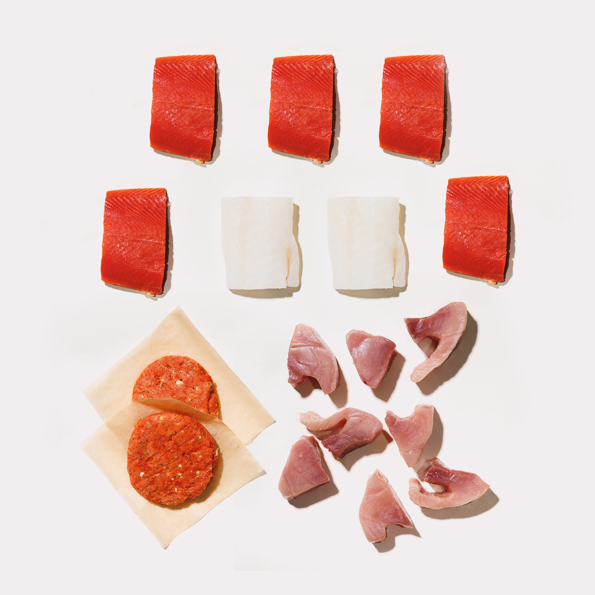 Raw sockeye salmon, pacific cod, and tuna medallions with two feta dill burgers on a white background