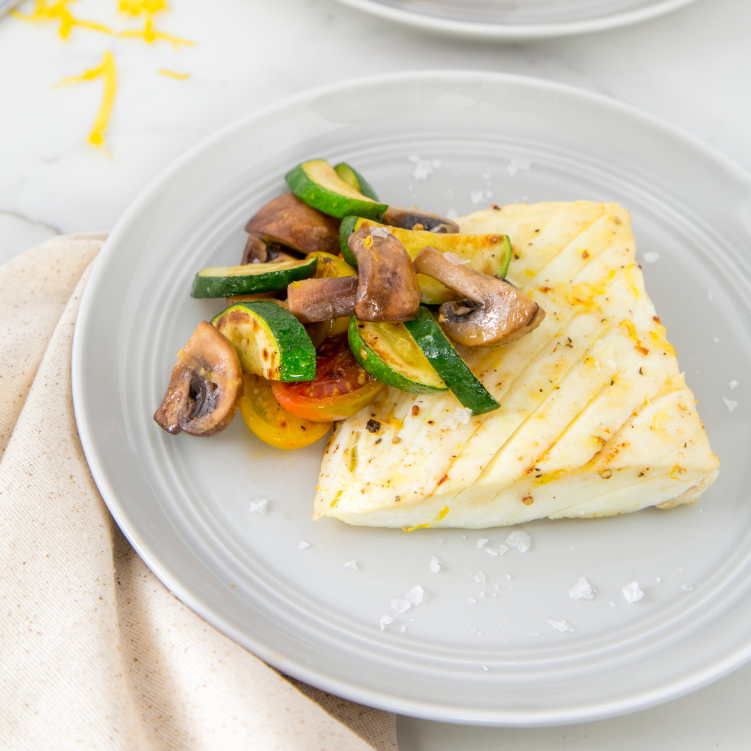 Plated dish of grilled halibut with zucchini, mushrooms, and tomatoes on a light background