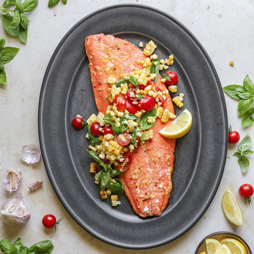 Cooked aalmon fillet with corn and tomato salad on a gray plate, surrounded by basil leaves and lemon wedges.s