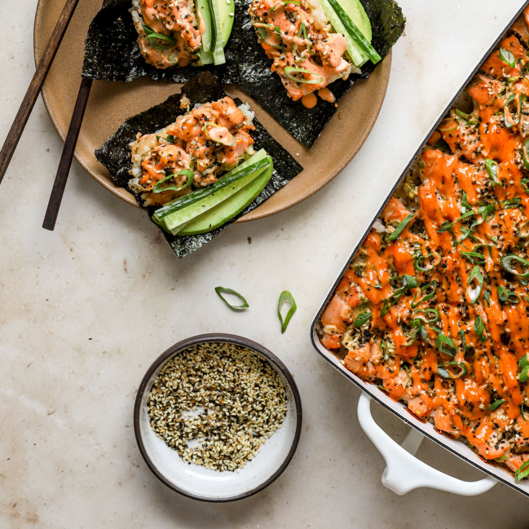 Sushi rolls with salmon and cucumber on a plate with chopsticks, sesame seeds, and a dish of Barnacle Foods Furikake seasoning.